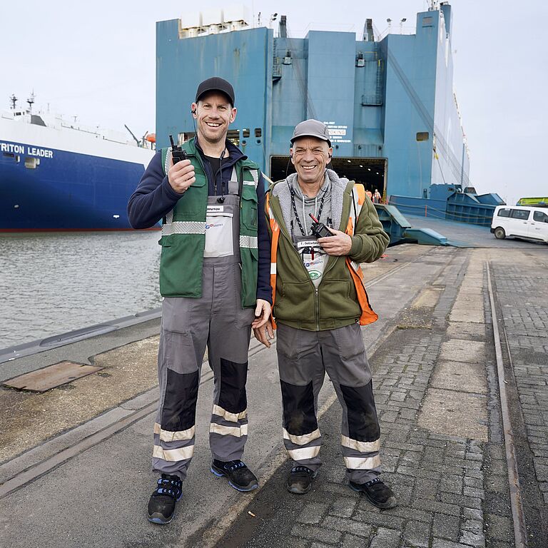 Zwei Hafenarbeiter mit Funkgeräten stehen vor Autotransportschiff im Hafen.
