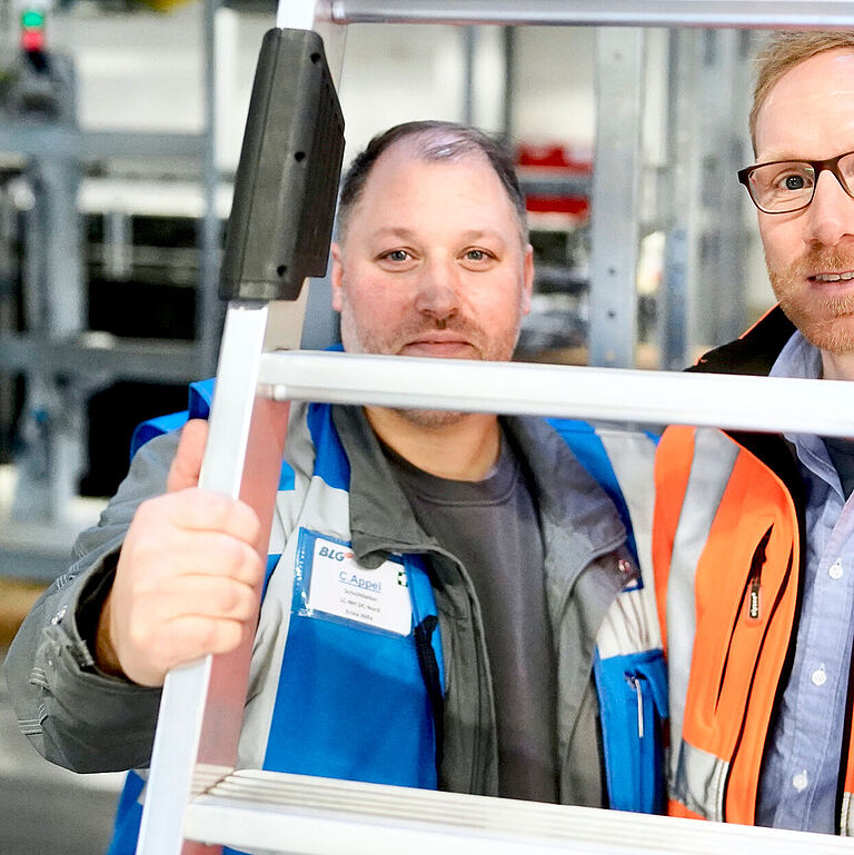 Schichtleiter Christian Appel und Thomas Menke, Leiter Operations Kommissionierung, schauen durch eine Leiter.