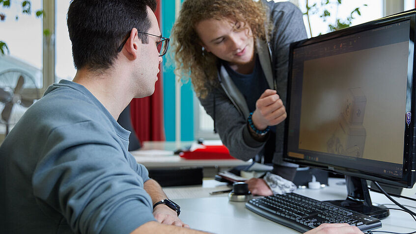 Tim sitzt an einem Tisch vor einem Computer, sein Kollege Arthur steht links von ihm und beugt sich zur Seite. Beide schauen auf den Bildschirm.