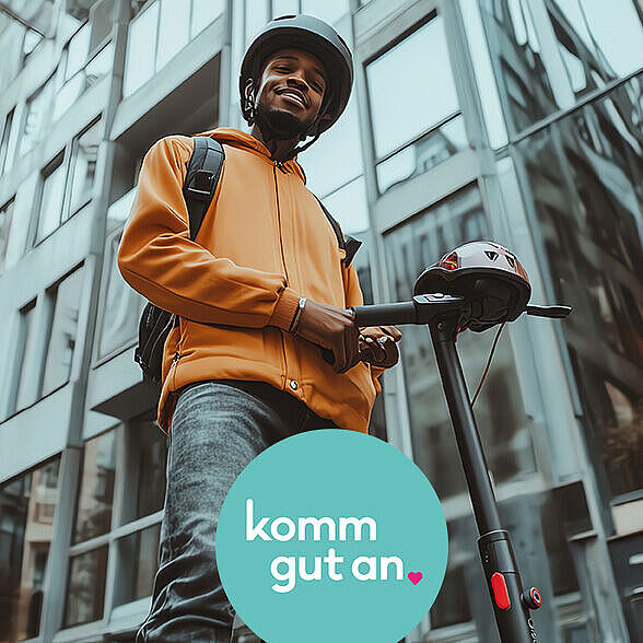 Ein junger Mann mit Helm und Rucksack steht auf einem E-Scooter in einer städtischen Umgebung mit modernen Glas- und Betongebäuden im Hintergrund. Er trägt eine orangefarbene Jacke, lächelt und blickt freundlich in die Kamera. Unten im Bild befindet sich ein rundes, türkisfarbenes Logo mit dem Text „komm gut an.“