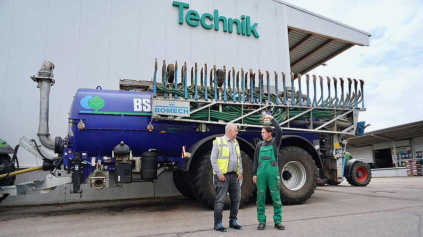 Ein Mann in einer Warnweste und ein Mann in grüner Arbeitslatzhose stehen vor einem Traktor mit einem Güllefass.