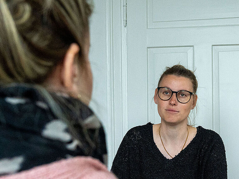 Gesprächssituation an einem kleinen Tisch: Eine Frau im rosa Pullover sitzt mit dem Rücken zur Kamera. Vor ihr sitzt eine junge Frau mit Brille im schwarzen Pulli und schaut sie freundlich an. Im Hintergrund ist eine weiße Holztür zu sehen. 