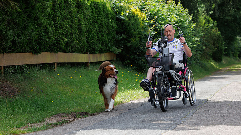 Dirk Boßmeyer trainiert mit seinem Hand-Bike, ein Rollstuhl-Zuggerät, das durch die Muskelkraft der Arme angetrieben wird. Der Schotterweg führt an einer grünen Hecke vorbei. Sein Hund begleitet ihn.