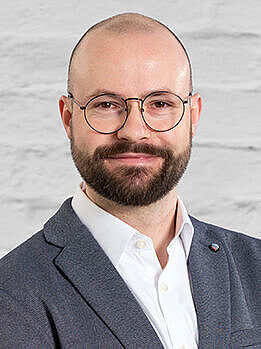 Porträt eines Mannes mit Brille, Bart und Anzug, vor hellem Hintergrund, mit verschränkten Armen. Es handelt sich um Nicolai Buchholz, ehrenamtliches Mitglied der BGHW-Selbstverwaltung für die Arbeitgeberseite.