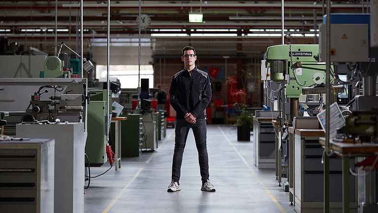Tim Mayer steht in einem Mittelgang in einer Werkstatt. Er hat die Hände vor dem Körper verschränkt, trägt Arbeitskleidung und eine Schutzbrille. Links und rechts von ihm stehen verschiedene Maschinen.
