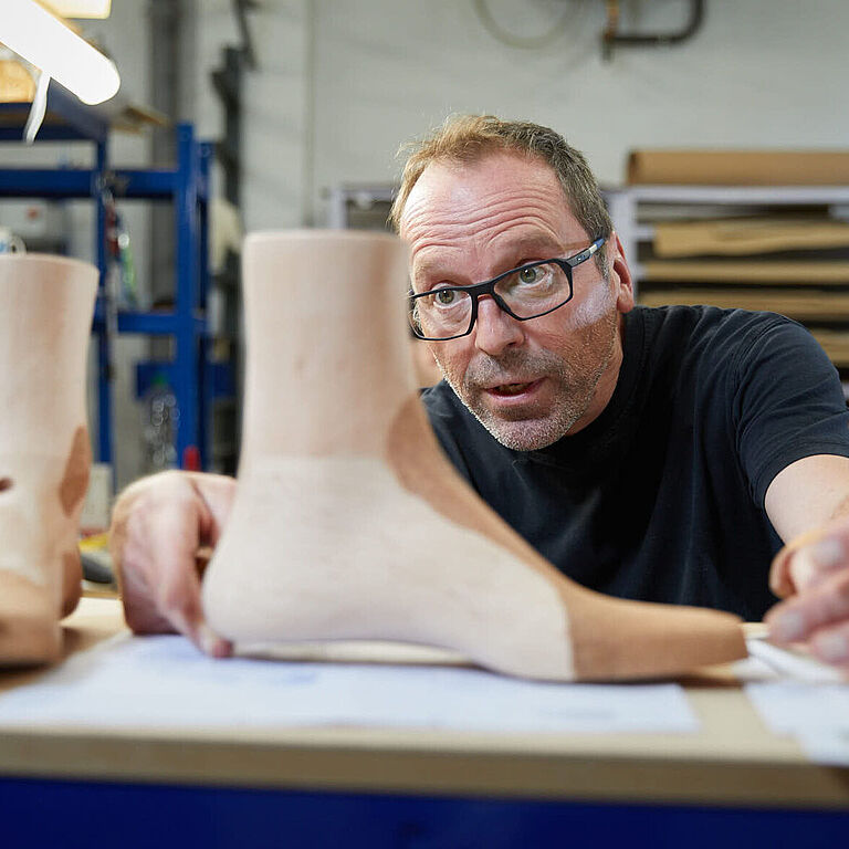 Ein Mann im schwarzen T-Shirt sitzt in einer Werkstatt und betrachtet zwei Holz-Leisten. Es sind die Muster, nach denen der Orthopädie-Schuhmachermeister die Maßschuhe fertigt. 