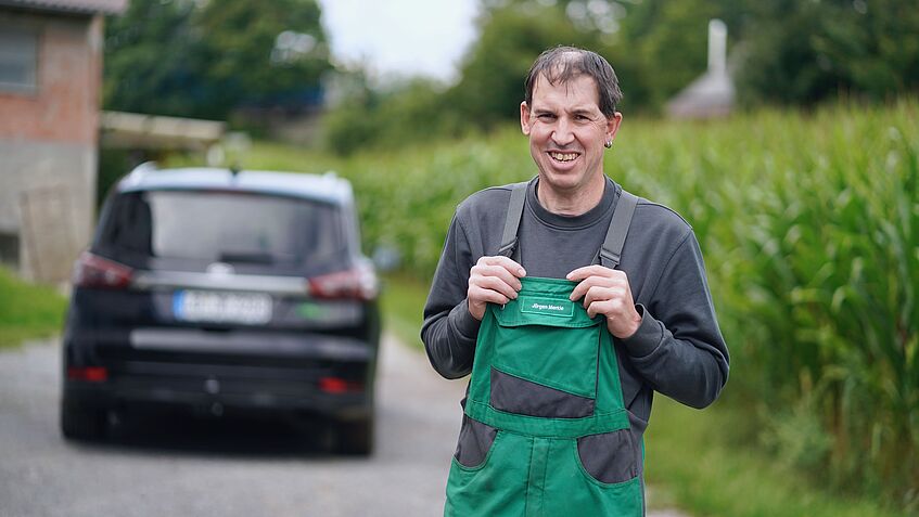 Ein Mann in grüner Arbeitslatzhose steht draußen vor einem Auto.