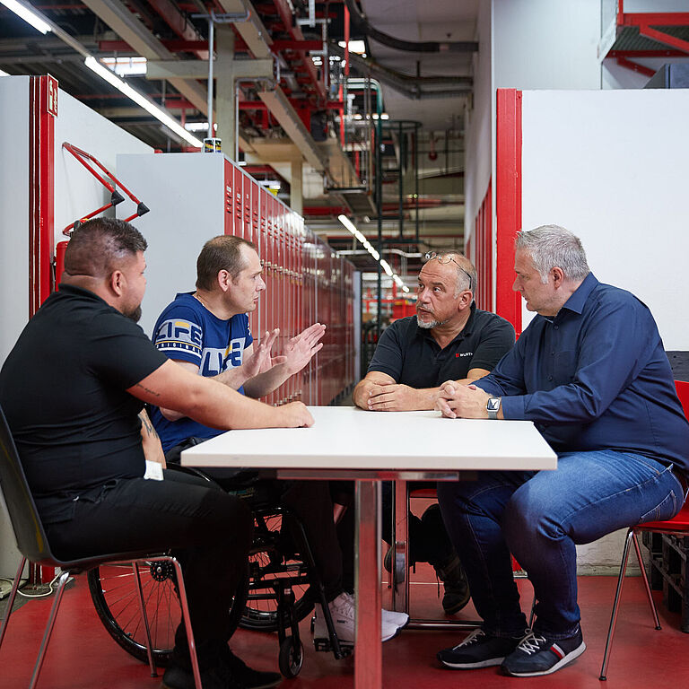 In einer Werkshalle der Firma Würth in Künzelsau sitzen vier Männer an einem Tisch und sprechen miteinander. Im Hintergrund sind Trennwände und Regale zu erkennen. Der zweite von links ist Johann Koch, der im Rollstuhl sitzt. 