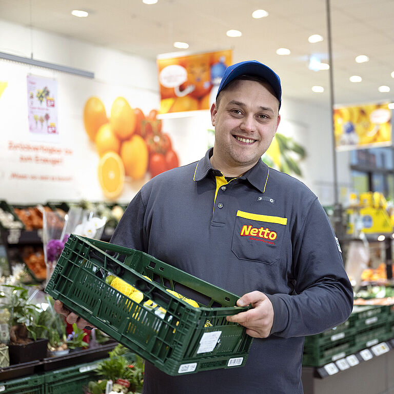 Dennis Zivanovic von Netto Marken-Discount in Arbeitskleidung und blauer Kappe lächelt in die Kamera, während er eine grüne Obst- und Gemüsekiste hält. Er steht in der Obst- und Gemüseabteilung des Supermarktes, umgeben von frischen Produkten und bunten Werbeplakaten im Hintergrund.