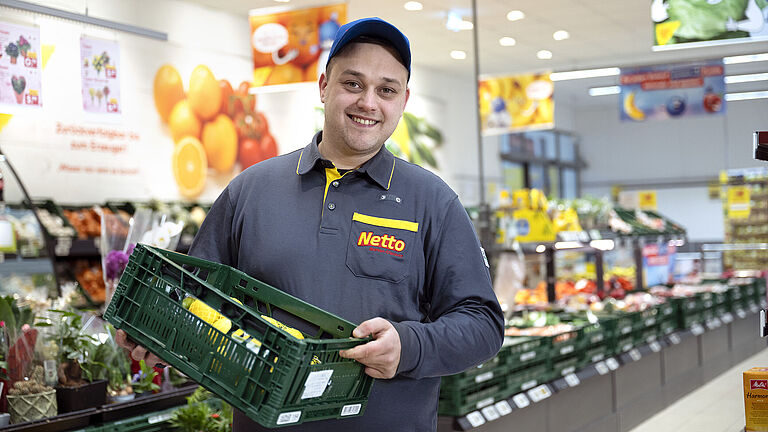 Dennis Zivanovic von Netto Marken-Discount in Arbeitskleidung und blauer Kappe lächelt in die Kamera, während er eine grüne Obst- und Gemüsekiste hält. Er steht in der Obst- und Gemüseabteilung des Supermarktes, umgeben von frischen Produkten und bunten Werbeplakaten im Hintergrund.