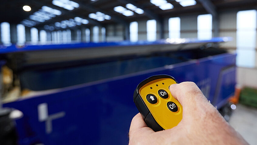 Eine linke Hand im Vordergrund hält eine gelbe Fernbedienung. Der Daumen drückt auf eine Taste. Im Hintergrund ist der Auflieger eines Lkw zu sehen. Er bewegt sich auf Tastendruck.