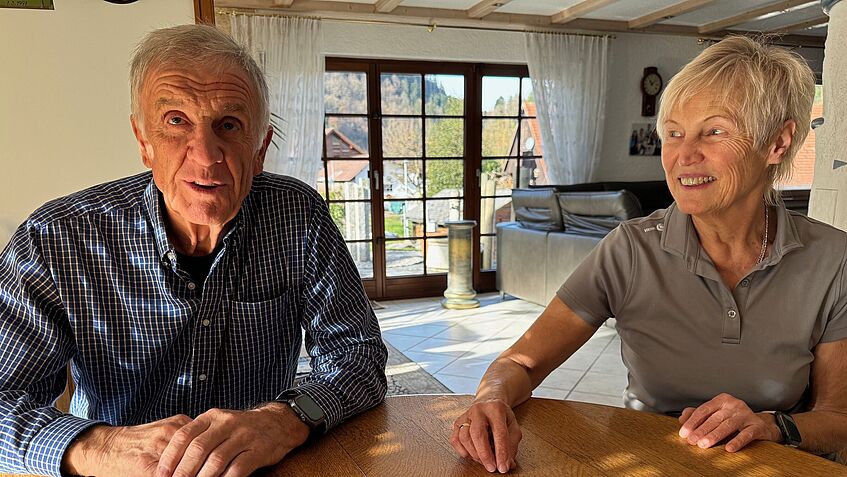 Die Großeltern von Lars sitzen an einem Holztisch und schauen freundlich in die Kamera.