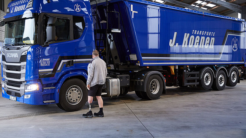 Ein Mann - in kurze Hosen - steht vor einem großen blauen Lkw. Er trägt links eine Unterschenkel-Prothese. Er ist auf dem Weg ins Lkw-Führerhaus, um seinem Job als Berufskraftfahrer zu erfüllen.