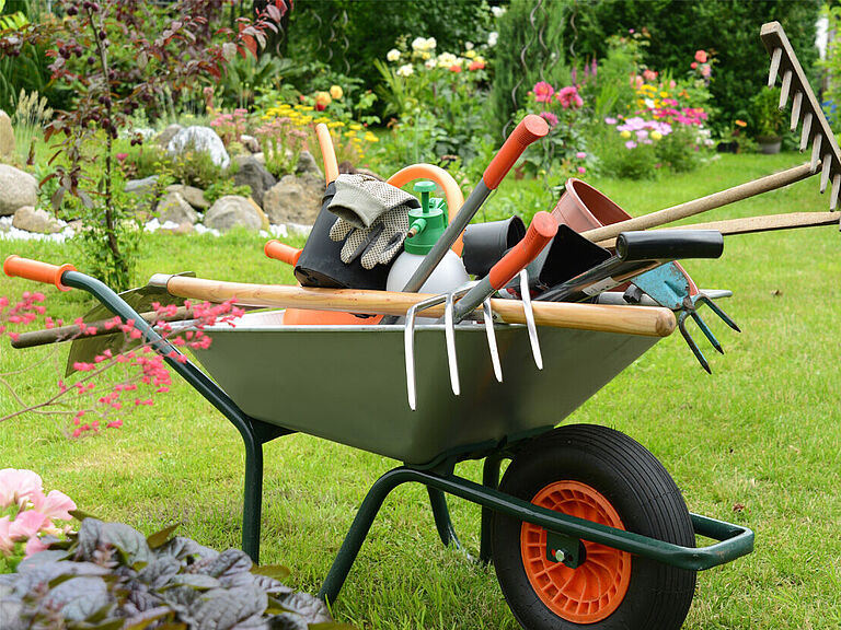 Schubkarren steht auf dem Rasen. Beladen mit Gartenwerkzeugen (wie Rechen, Spaten, Gartenhandschuhen etc.), die unordentlich übereinander liegen. 