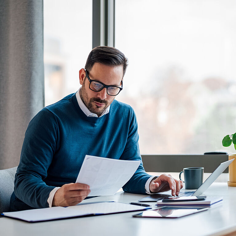 Mann liest ein Dokument und tippt auf einem Laptop, an einem Schreibtisch mit Büromaterial.