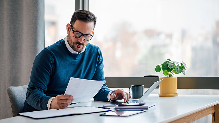 Mann liest ein Dokument und tippt auf einem Laptop, an einem Schreibtisch mit Büromaterial.