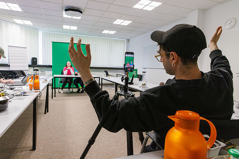 Dreharbeiten: Im Vordergrund hebt ein Mann die Hände, im Hintergrund drehen junge Menschen vor einem Greenscreen