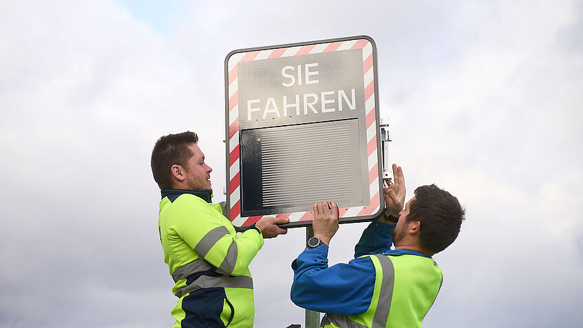 Zwei Mitarbeiter der Firma TST auf einer Leiter. Sie montieren das Display des Geschwindigkeitsmessgerätes.