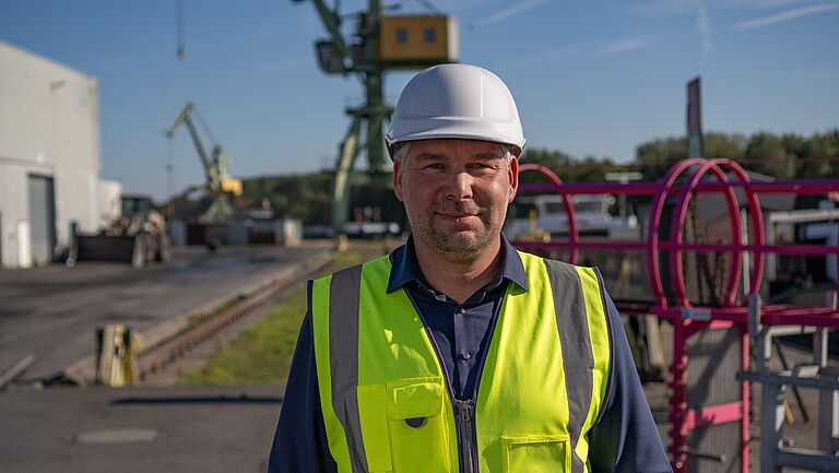 Jörg Hörstemeier, der Technische Leiter der Stadthafen Lünen GmbH, guckt freundlich in die Kamera. Er trägt einen Helm und eine Warnweste. Hinter ihm ist ein Teil des Hafens zu sehen.