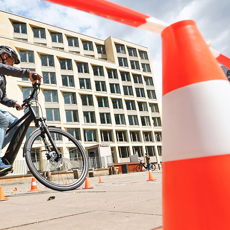 Ein Platz mit einer leuchtend rot-weiße Pylone im Vordergrund, darüber weht ein rot-weißes Flatterband. Ein Zweiradfahrer umkurvt Pylonen. Im Hintergrund sind mehrstöckige Häuserfassaden zu sehen. 