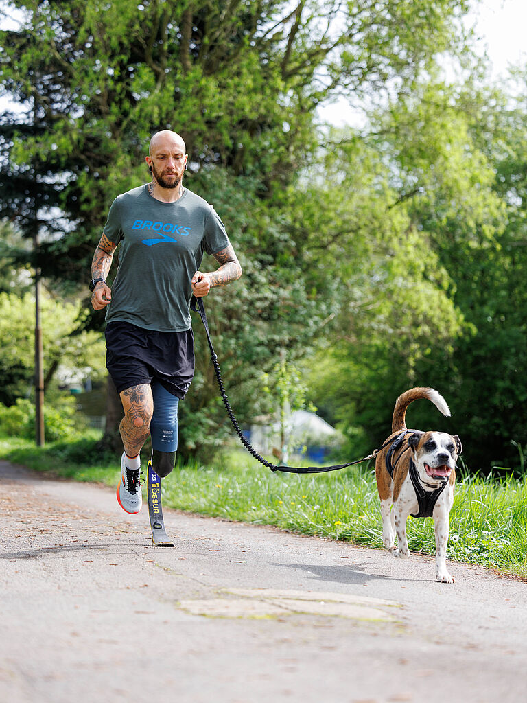 Para-Triathlet Kim Cremer trainiert gerne mit seiner Hündin Nikita. Hier läuft er über einen Asphaltweg im Grünen, sie vorneweg mit heraushängender Zunge. 