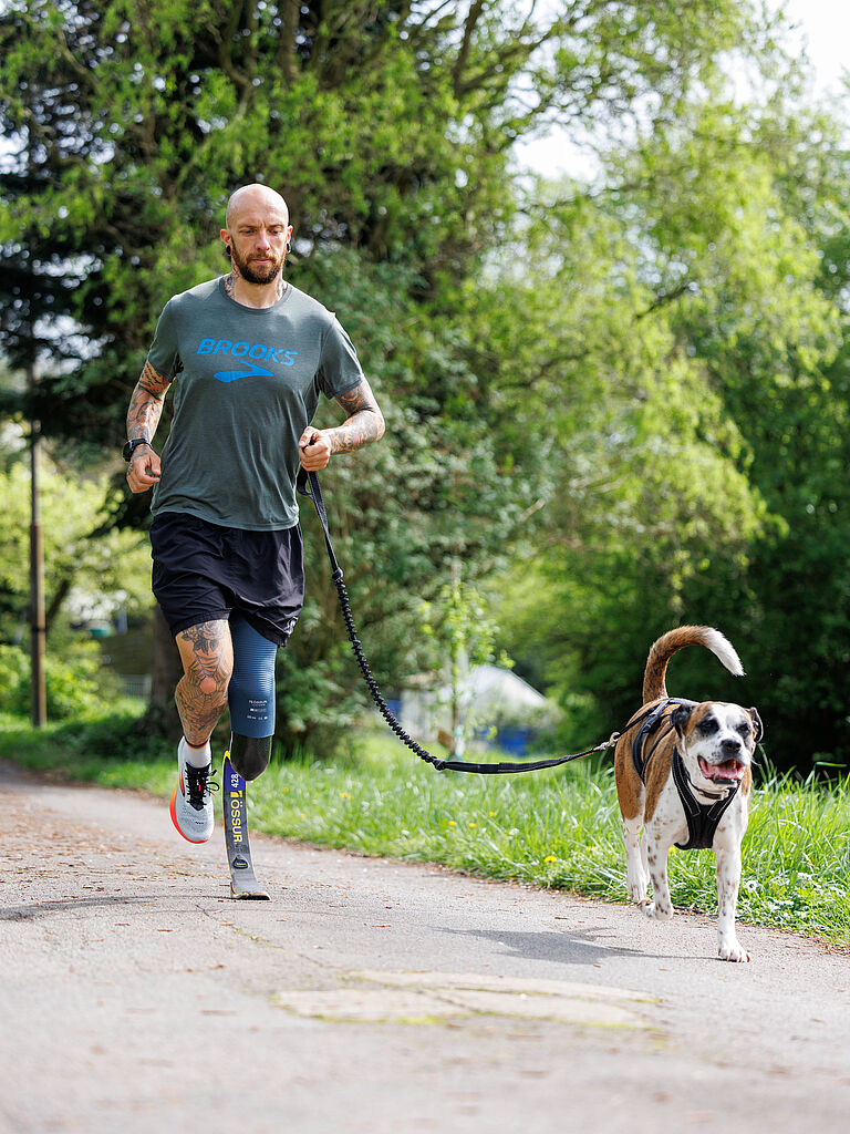 Para-Triathlet Kim Cremer trainiert gerne mit seiner Hündin Nikita. Hier läuft er über einen Asphaltweg im Grünen, sie vorneweg mit heraushängender Zunge. 