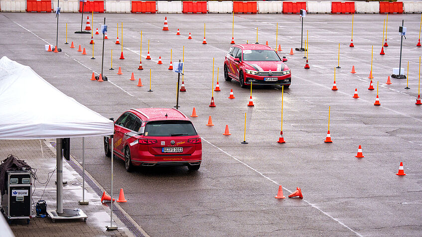 Blick von oben: Zwei rote Pkw fahren in einem abgesteckten Parcours. Links steht ein Zelt als Regenschutz für das Orga-Team. Die Bahnen sind durch orangefarbene leuchtende Pylonen abgesteckt. Die Fahrzeuge kommen sich in sicherer Entfernung entgegen. Zwei Studienteilnehmende werden parallel getestet. 