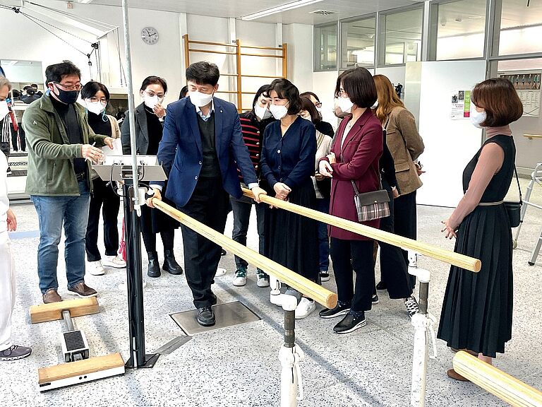 Eine Besuchergruppe aus Korea informiert sich in der BG-Klinik Ludwigshafen. Dabei schauen sie sich auch ein Gerät an, das ähnlich wie ein Stufenbarren ist 