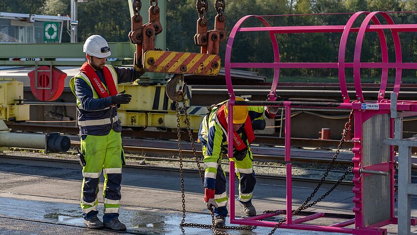 Zwei Hafenmitarbeiter in Schutzkleidung befestigen ein mobiles Leiterkonstrukt an einem Kran.