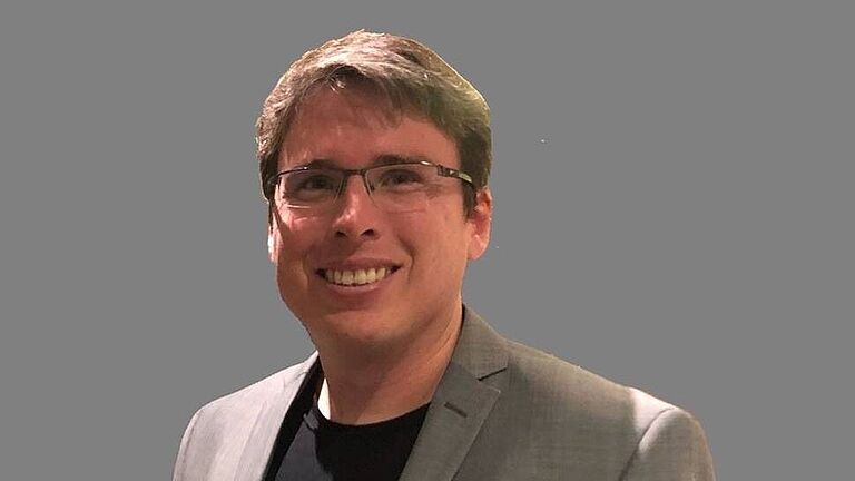 Lächelnder Mann mit Brille, grauem Sakko und schwarzem Shirt vor grauem Hintergrund