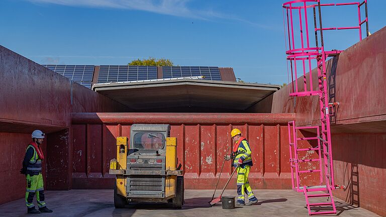 Zwei Mitarbeiter in Schutzkleidung säubern die Ladefläche von einem Schiffsladeraum, an der rechten Seitenwand ist eine mobile Einstiegsleiter zu sehen. 