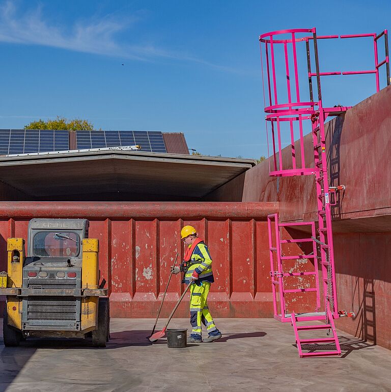 Zwei Mitarbeiter in Schutzkleidung säubern die Ladefläche von einem Schiffsladeraum, an der rechten Seitenwand ist eine mobile Einstiegsleiter zu sehen. 