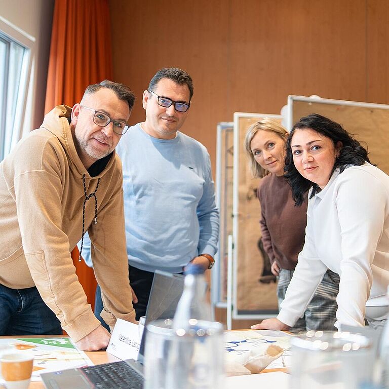 Vier Personen stehen in einem Seminarraum zusammen und schauen in die Kamera