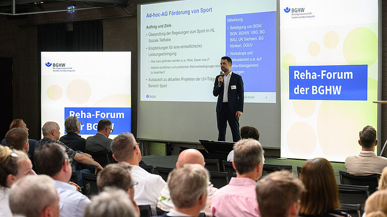 Joscha Schwarzwälder, DGDGUV-Referatsleiter Heilbehandlung und Teilhabe, steht auf der Bühne des Reha-Forums, links von im sitzen die Teilnehmenden. Im Hintergrund erstrahlen seine Präsentation und die Logos der BGHW auf Display-Wänden. 