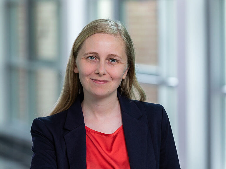 Porträt einer lächelnden Frau mit langen hellbraunen Haaren in einem dunklen Blazer und roter Bluse vor einem unscharfen Innenraumhintergrund.