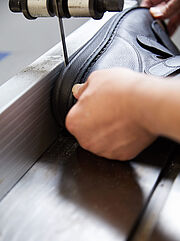 Ein Schuh, der von zwei Händen gehalten wird, liegt auf einem Metallband. Über ihm hängt ein schmales Bohrmesser, das die Schuhsohle längs auftrennt. 