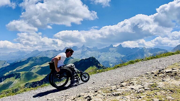 Christoph Weis fährt fröhlich mit einem Handbike einen Berg hoch. Im Vorder- und im Hintergrund: Berglandschaft, Natur pur. 