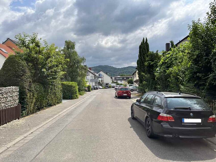 Wohnstraße mit parkenden Autos und Häusern, Hügel im Hintergrund.