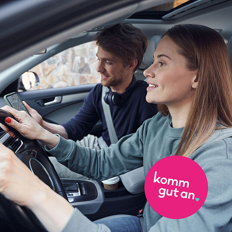 Zwei angeschnallte Personen sitzen in einem Auto. Die Person am Steuer blickt nach vorn, die Person auf dem Beifahrersitz hält ein Smartphone. Auf dem Bild ist der Schriftzug „komm gut an“ eingeblendet.