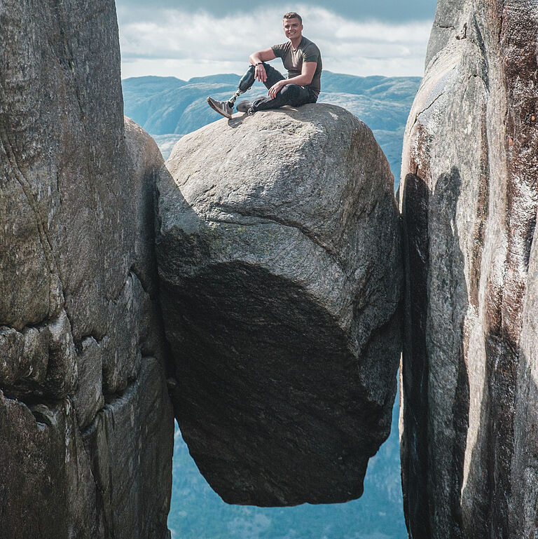 Thomas Müller, ein BGHW-Versicherter aus Köln, dessen rechter Unterschenkel nach einem Unfall amputiert werden musste, sitzt auf einem großen felsigen Stein, der zwischen zwei Felsen on der Luft hängt. n hängt und schaut in die Kamera. Er schaut freundlich in die Kamera.