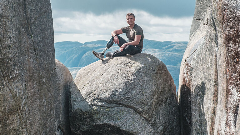 Thomas Müller, ein BGHW-Versicherter aus Köln, dessen rechter Unterschenkel nach einem Unfall amputiert werden musste, sitzt auf einem großen felsigen Stein, der zwischen zwei Felsen on der Luft hängt. n hängt und schaut in die Kamera. Er schaut freundlich in die Kamera.
