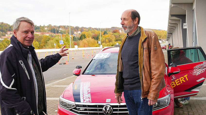 Zwei Männer stehen vor einem roten Pkw. Der Mann links, Professor Matthias Graw, zeigt auf das Fahrzeug und schaut in die Kamera. Der Mann rechts, Professor Wolfgang Fastenmeier, schaut auf die andere Person. 