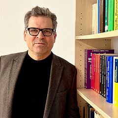 Nikolaus Lange schaut freundlich in die Kamera. Er trägt eine Brille, einen schwarzen Pullover und eine braune Jacke und steht vor einem Bücherregal.