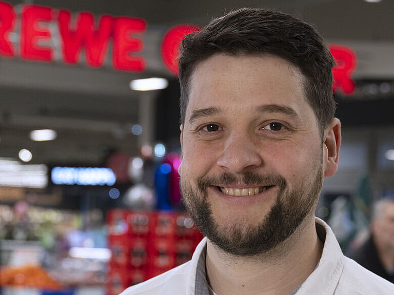 Marktleiter Moritz Breske in weißem Hemd mit Namensschild steht lächelnd in einem gut beleuchteten REWE-Markt vor einem bunten Hintergrund aus Regalen, Obstständen und Schildern. Das Schild über ihm trägt den Schriftzug ‚REWE CENTER‘.