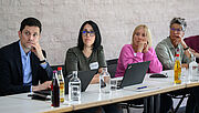Drei Frauen und ein Mann sitzen an weißen Tischen nebeneinander in einem Workshop Vor ihnen liegen Unterlagen, Laptops und Getränke stehen auf den Tischen. Der Mann sitzt links, die Frauen rechts nebeneinander. Sie alle schauen aufmerksam nach links. 