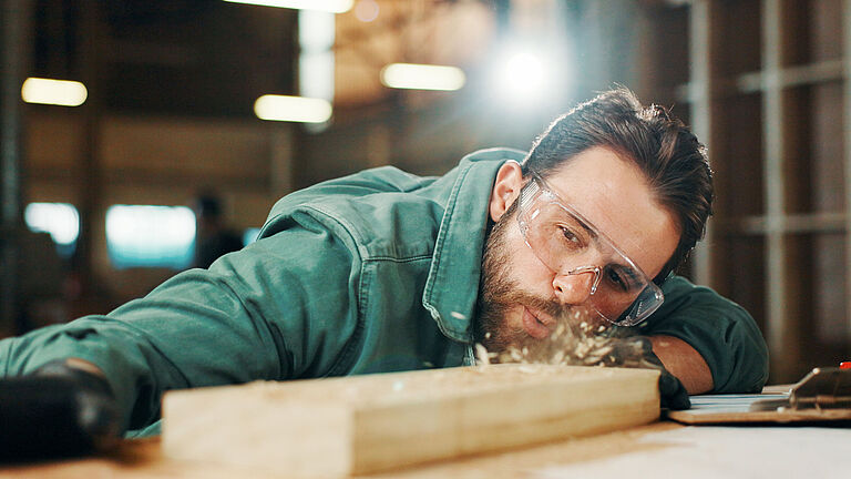 Das Bild zeigt einen Tischler mit Schutzbrille, der Späne von einem Holzstück pustet. 
