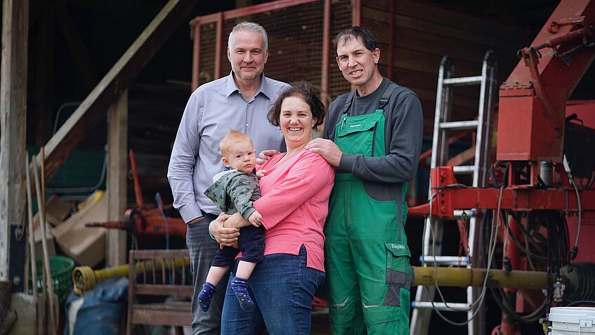 Zwei Männer und eine Frau mit Kleinkind auf dem Arm stehen draußen, im Hintergrund Bauernhof-Ambiente mit Leiter und Landmaschine.