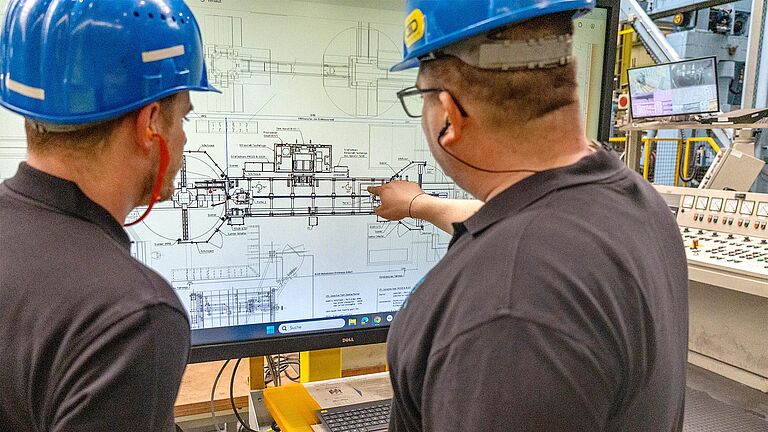 Zwei Männer mit blauen Schutzhelmen schauen auf einen Computerbildschirm.