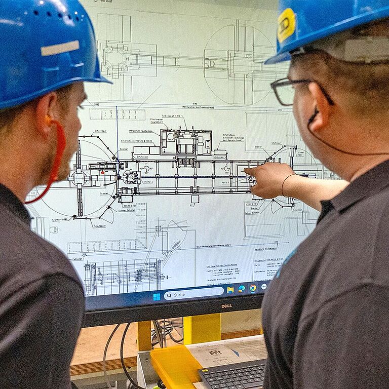 Zwei Männer mit blauen Schutzhelmen schauen auf einen Computerbildschirm.