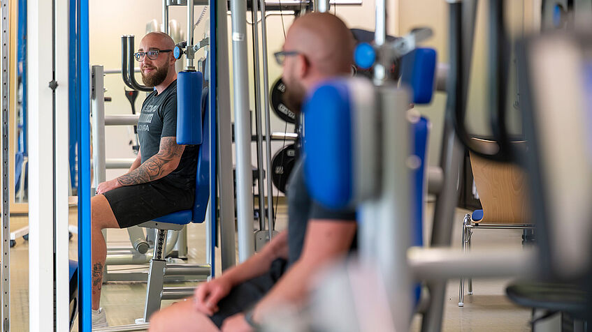 Mortz Sontheimer sitzt in einem Fitnessraum an einem Kräftigungsgerät. Er ist seitlich von hinten zu sehen, gegenüber sieht man ihn frontal im Spiegelbild.